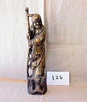 Front left angle of the hand-carved wooden statue of an old man holding a staff, showing detailed facial features and robe folds.