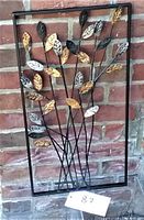 Photo of metal wall art showing rectangular frame with metal leaves in gold, silver, and black colors mounted on thin metal rods against a brick wall.