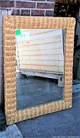 Mirror standing outdoors leaning against a white metal vertical door and brick wall. The thick woven straw frame is visible.