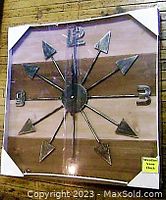 Front view of large square wall clock with metal weathervane style hands and numbers 3, 6, 9, 12 on wooden plank face.