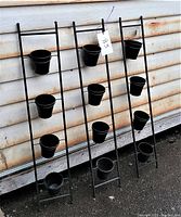 Three vertical black metal garden ladders with four black removable metal flower pots on each ladder. Total 12 pots. Photo shows front view of all three ladders leaning against a wall.