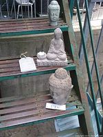 Photo of weathered concrete Buddha bust with chipped nose, ceramic Buddha figurine, and small glazed Buddha head on metal stairs
