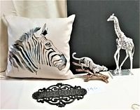 Zebra cushion showing full design of zebra head next to the leopard and giraffe resin figures and metal welcome sign on a white surface.