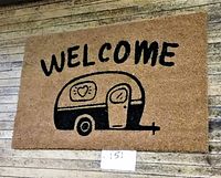 Full view of rectangular brown coir welcome mat with black print of old-style trailer and 'WELCOME' text, displayed hanging on a wood wall.