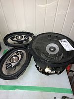 Three car speakers displayed on a table, showing one large round speaker with transparent cover and two smaller oval metal cone speakers.