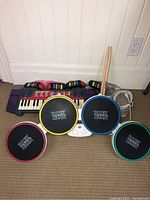 Rock Band drum set with four color-coded drum pads and two wooden drumsticks placed beside an electronic keyboard with multi-colored keys and controls.
