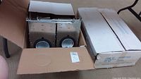 Two opened cardboard boxes, each containing four recessed light fixtures with metal backs and white sockets visible.
