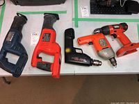 Four power tools on a white table: two reciprocating saws on the left, two drills on the right.