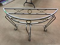 Front view of a demilune metal table base showing the half-moon shape with decorative circular and X design elements, curled legs, metallic finish.