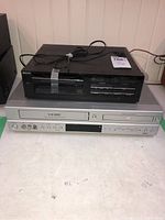 Philips black VCR player stacked on top of Viore silver VCR player with visible buttons and tape slots