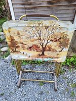 Four vintage metal TV trays with scenic pastoral artwork stacked in a gold metal holder with a handle, displayed outdoors on gravel ground near a wooden wall.