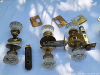 Four sets of antique glass door knob lock hardware with brass plates and latches laid out on a white surface under some shade.