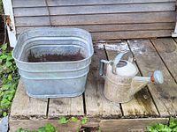 Side view of galvanized watering can and square washtub on wooden surface outdoors, showing condition and relative size of items.