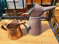 Overall view of two copper pouring cans placed on a wooden table; large can stacked on the smaller can showing size difference and shape.