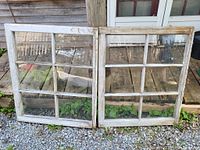 Pair of antique whitewashed pine window frames with all glass panes intact, set outside on gravel and a wooden porch showing rustic weathering on the wood.