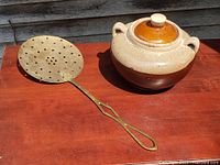 Photo showing ceramic bean pot with lid and brass skimmer on wooden surface