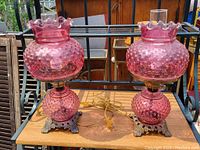 Both cranberry hobnail glass table lamps side by side on shelf