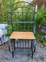 Front view of metal bakers rack showing decorative metal frame, wood-like shelf, and wine holder rack below the shelf.
