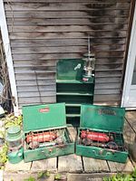 Lot arranged outside against siding showing two green Coleman lanterns on the wooden surface, two green Coleman portable stoves with red tanks in front of a green metal shelf unit with three shelves.