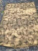 Full view of the rectangular Oriental style rug laid out on a gravel surface, showing the beige background with dark brown ornamental scroll and vine design.