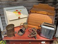 Photo showing six vintage kitchen items including two bread boxes (one cream colored with floral design, one wooden), chrome toaster, metal sifter, metal scoop, and ceramic mug on a wooden table.