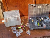 Photo of fishing tackle lot arranged on wooden surface including the two plastic tackle boxes (grey and white translucent) and various fishing lures, both hard plastic and soft plastic swimbaits, plus small packs of lure tails and accessories.