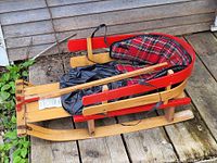 Photo of wooden child's sleigh with red painted accents, natural wood finish, padded cushion in red plaid fabric, and black leather strap.
