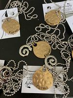 Seven necklaces laid out showing the gold-tone hammered round pendants with 'Karma' text and smaller silver spiral charms on silver ball chains, all with tags.