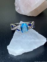 Close-up of aquamarine ring with clear cubic zirconia swirls and dark blue small stones on silver-tone band, placed on white and pink stones.