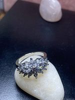 Close-up photo of a silver ring displaying a floral cluster design with multiple clear cubic zirconia stones on a white stone background.