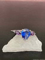 Front view of the ring showing the blue tear-shaped central gem with purple oval side gems on a silver band, placed on a piece of white quartz.