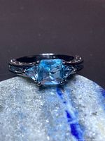 Front-on view of the blue topaz ring placed on a mottled stone surface, showing the square-cut center gemstone and the two smaller side stones with the dark metal band and detailing.