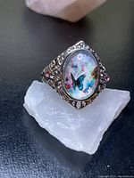 Front angled view of the butterfly ring showing the oval center with butterfly design, pink side stones, and filigree band detail.