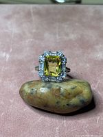 Front-close up photo of the citrine gemstone ring resting on a smooth brown stone, highlighting the rectangular yellow citrine center and surrounding clear stones in a halo.