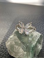 Close-up of silver-tone ring with clear cubic zirconia stones forming butterfly shape, placed on a bluish-gray textured surface.
