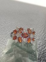 Front angle view highlighting two orange flower designs with clear CZ accents on silver ring band placed on green crystal base and black textured background.