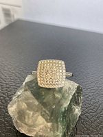 Ring displayed on a greenish crystal rock showing the pavé setting of cubic zirconia stones and the square cluster design.