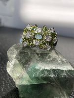 Front view of a silver-tone ring with two clusters of deep green flower-shaped stones surrounded by clear cubic zirconia stones, shown on a green crystal.