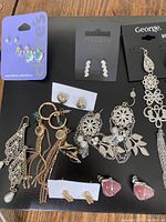 Lot of assorted earrings displayed on black surface including brands Pilgrim, George, Claires, with various designs such as dangle chains, filigree, and colorful stones.