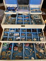 Opened three-tier white jewelry box showing top and middle trays with compartments filled with necklaces, bracelets, brooches