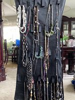 Necklaces displayed hanging vertically on a black strap organizer, showing various designs including bead strands, chains and pendant necklaces.