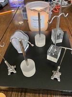 Five jewelry holders arranged on a black surface near a lit candle. Varied bases including metallic Eiffel Towers and acrylic blocks, all with metal T-shaped holders.