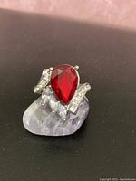 Front left angle close-up showing large pear-shaped red gem with white accent stones on a silver-tone setting, ring placed on a light purple stone.