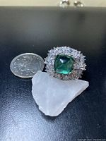 Front view of large ring showing deep green main stone surrounded by clear cubic zirconia floral clusters next to a quarter for scale.