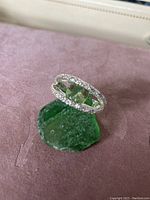 Ring displayed on a green rough stone on soft pink fabric background, showing the rectangular light green gemstone and clear crystals on silver band.