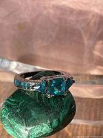 Close-up photo of the sterling silver ring with blue-green square center stone on green textured surface.