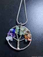 Close-up photo of silver Tree of Life pendant necklace with wire tree design and multi-colored stone chips on black background.