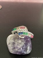 Silver-tone ring with rows of square-cut blue, green, pink, and yellow gems along with many small white gems in a wavy band design, displayed on a purple stone.