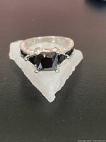 Front close-up of silver-tone ring with square black gemstone centerpiece on a white crystalline base.