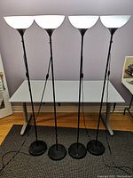 Four identical black floor torchiere lamps with white shades lined up on a table against a gray wall.
